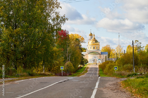 Asphalt road leading to the village with a beautiful white stone Orthodox Church with a yellow dome among nature and green trees. Natural landscape with greenery on a summer, autumn or spring day © keleny