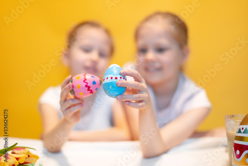 Cute little sisters with colorful eggs sitting at table with palette and Easter symbols against yellow background