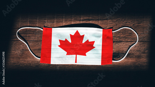 Canada National Flag at medical, surgical, protection mask on black wooden background. Coronavirus Covid–19, Prevent infection, illness or flu concept photo. State of Emergency, Lockdown
