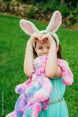 Cute little girl wearing bunny ears on Easter day plays with rabbit toy holding it in her hands and showing shape of heart on grass in backyard.