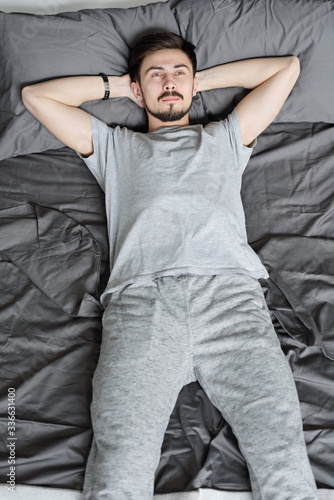 Pensive young bearded man in homewear lying with hands behind head on bed while relaxing at quarantine