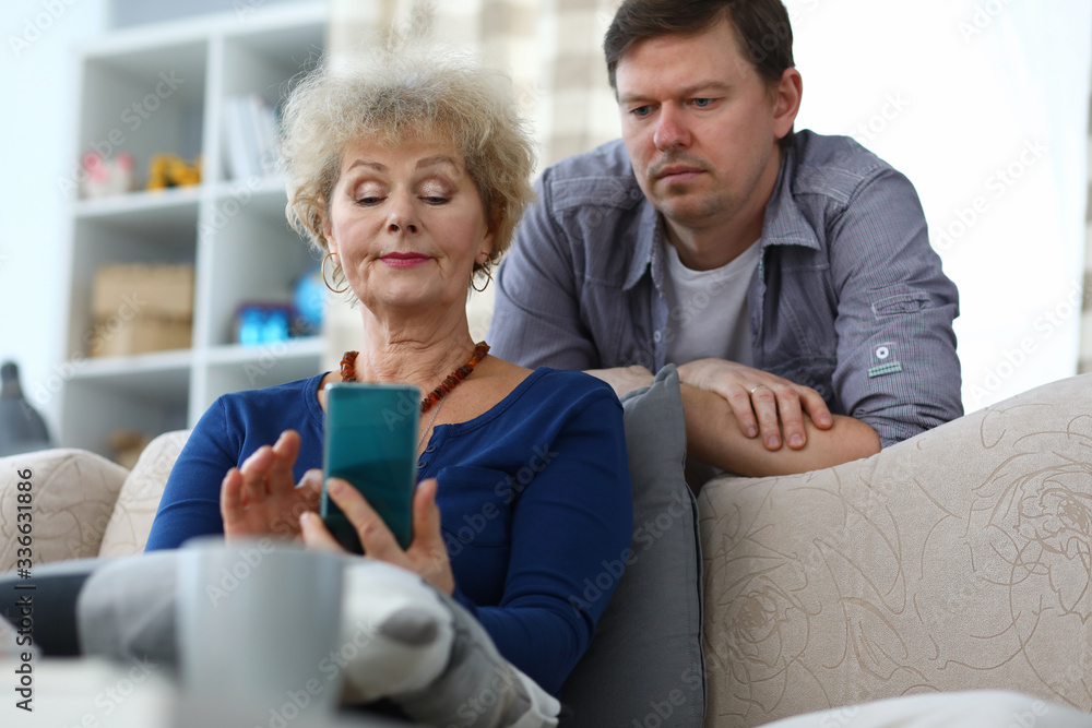 Portrait of senior woman using mobile phone for entertainment. Middle-aged male son standing behind couch. Cozy home interior. Fun family time and technology concept Portrait of senior woman using mobile phone for entertainment. Middle-aged male son standing behind couch. Cozy home interior. Fun family time and technology concept