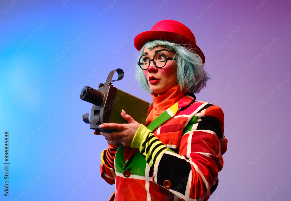 Obraz premium Close-up portrait of a clown girl with make-up, blue hair, a red hat, a colored checkered jacket and glasses.