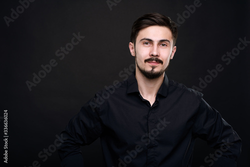Portrait of content handsome young man with mustache and beard standing against black background