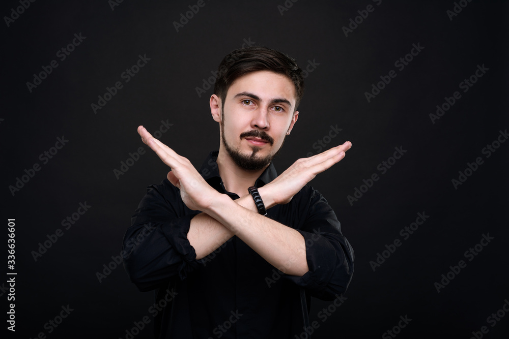 Portrait of young bearded man standing against black background and crossing arms while making stop gesture Portrait of young bearded man standing against black background and crossing arms while making stop gesture
