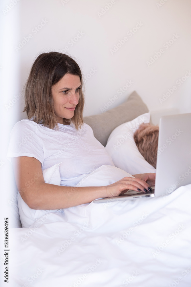 A beautiful woman works at a computer in her cozy white bed while her pink haired friend sleeps on a pillow A beautiful woman works at a computer in her cozy white bed while her pink haired friend sleeps on a pillow