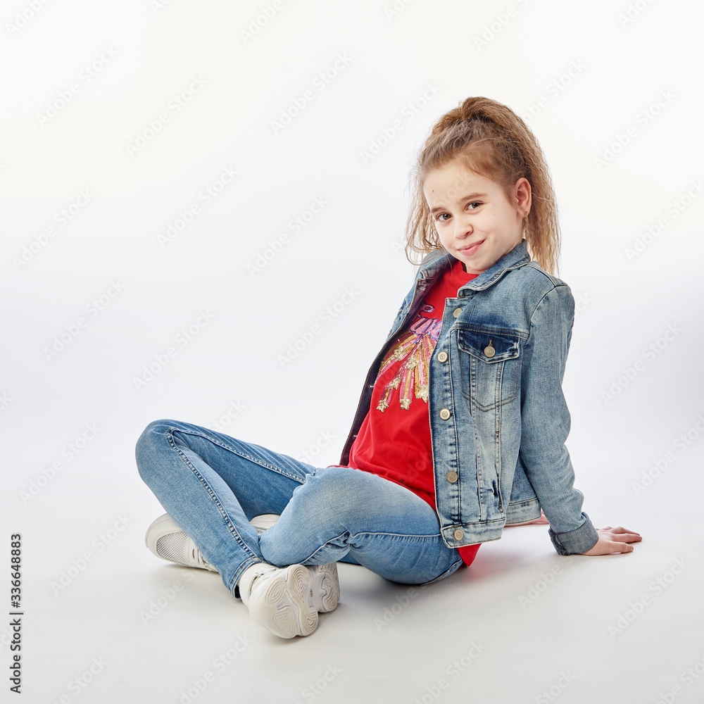Young girl wearing denim jacket, red shirt, jeans and white sneakers sitting cross-legged on floor Young girl wearing denim jacket, red shirt, jeans and white sneakers sitting cross-legged on floor