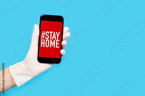 A hand in a glove holds a phone with the words Stay at home isolated on a blue background. 