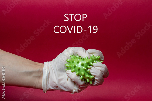 stop covid-19 hand in a medical glove tightly compresses a coronavirus on a red background with text