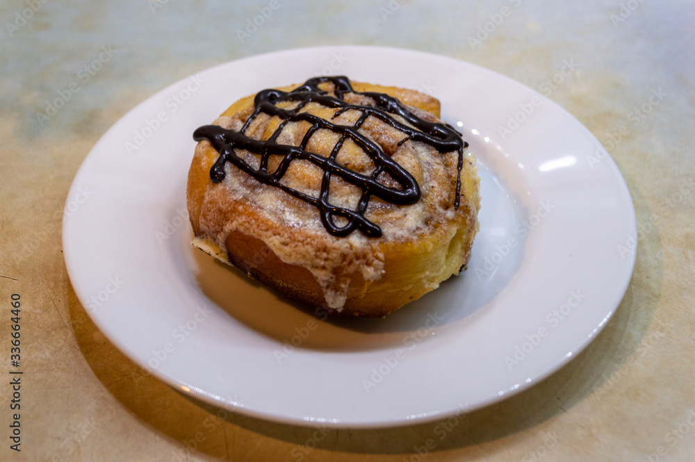 Cinnabon. Cinnamon roll decorated with chocolate on a white plate on blue and yellow kitchen table background Cinnabon. Cinnamon roll decorated with chocolate on a white plate on blue and yellow kitchen table background
