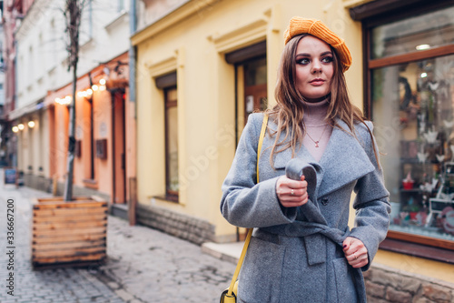 Portrait of stylish young woman wearing yellow beret tying coat holding purse on street. Spring fashion accessories
