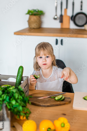 Cute kid, little girl cutting cucumber for green salad at kitchen