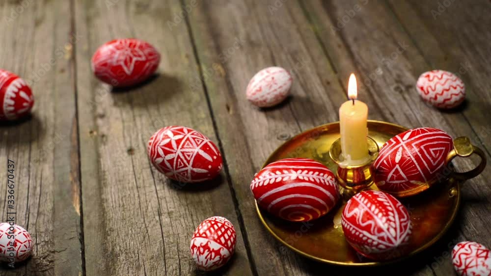Red easter eggs with folk Ukrainian pattern lay around and on candlestick with burning candle. And man rotate one egg on vintage boards background. Ukrainian traditional eggs pisanka and krashanka. Red easter eggs with folk Ukrainian pattern lay around and on candlestick with burning candle. And man rotate one egg on vintage boards background. Ukrainian traditional eggs pisanka and krashanka.