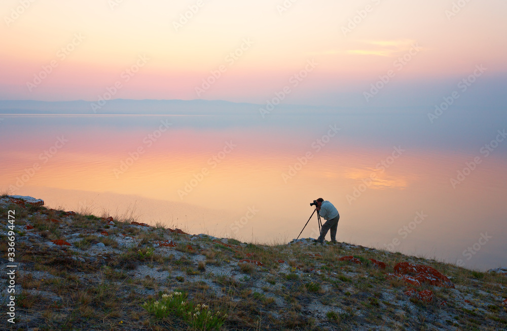 Baikal Lake on June. Silhouette of a tourist with a camera on a tripod on the rocky shore of Olkhon Island on the background of a beautiful sunset over the Small Sea. Photo tourism and travel concept