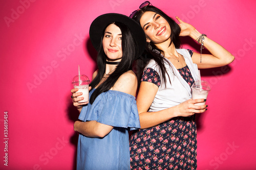 Two caucasian brunette hipster woman in casual stylish outfit having fun drinking milkshakes with straws. They standing on a bright pink background. Cheerful, happy emotions