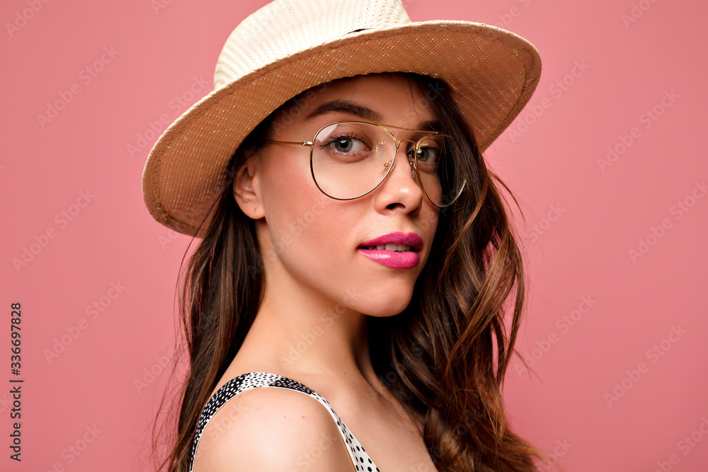 Close up indoor portrait of adorable lovely woman wearing stylish spectaculars and summer hat with pink lipstick posing over pink background Close up indoor portrait of adorable lovely woman wearing stylish spectaculars and summer hat with pink lipstick posing over pink background