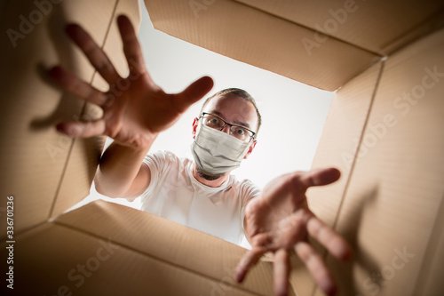 Young man opening the huge postal package wearing protective face mask. Male model on top of cardboard box. Food and goods contactless delivery during coronavirus quarantine for isolated people.