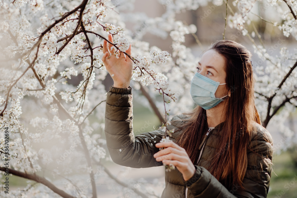 A girl with a medical mask on her face, allergic to flowers, stands in the garden of flowering cherry trees on a warm spring spring