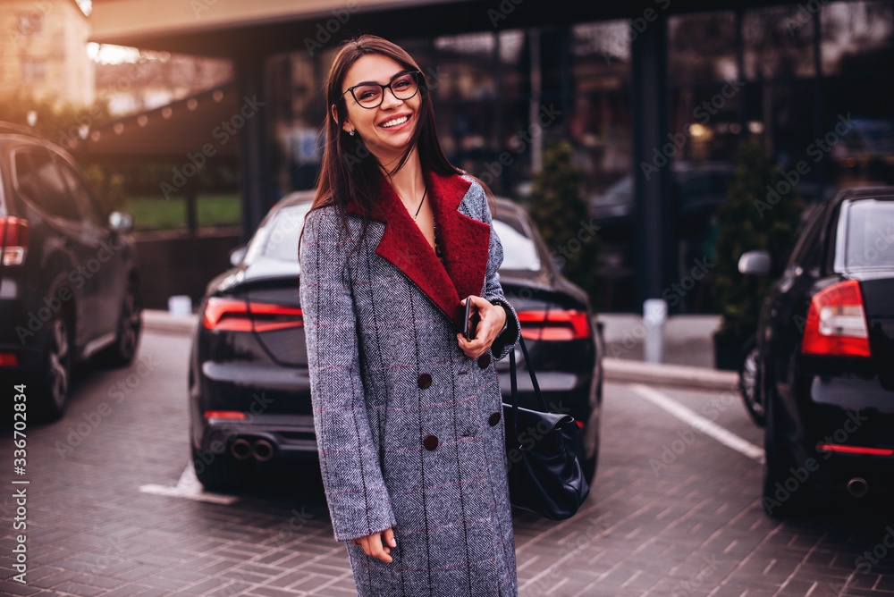 Smiling business woman with a smartphone standing near her car. Woman in eyeglasses with bag in hand