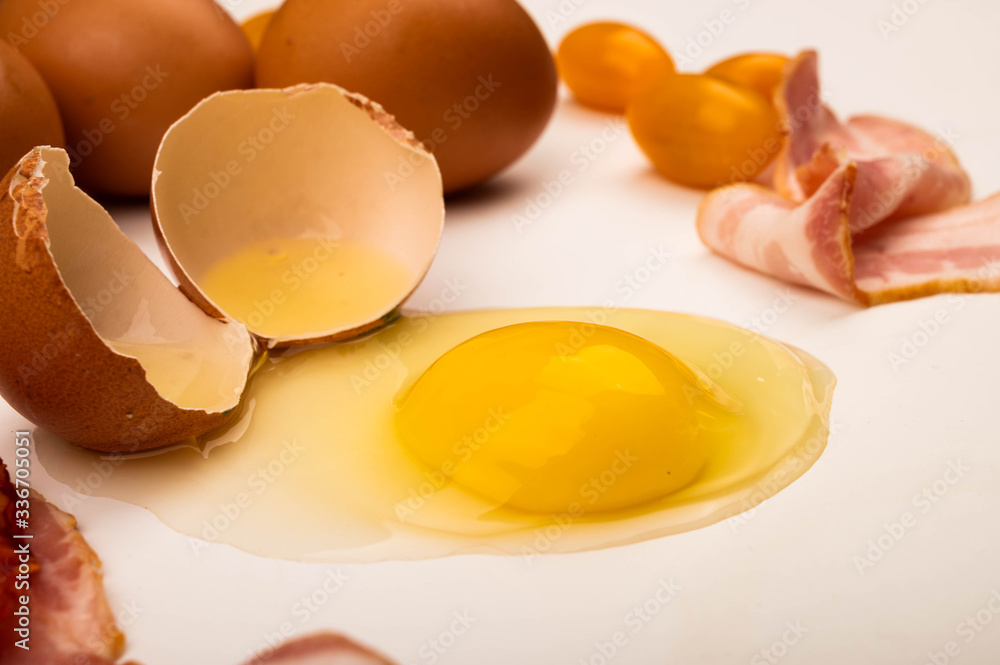 Broken chicken egg, chicken eggs, bacon slices and tomatoes on a white background. Close up. Broken chicken egg, chicken eggs, bacon slices and tomatoes on a white background. Close up.