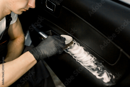 Cropped image of washing car interior by a soft brush with foam. Young male worker in black protective gloves cleaning car door inside with foam and brush