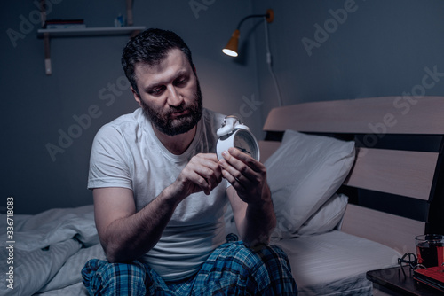 Tablou pe pânză A lonely bearded dark-haired man sitting on the bed at night sets the alarm clock
