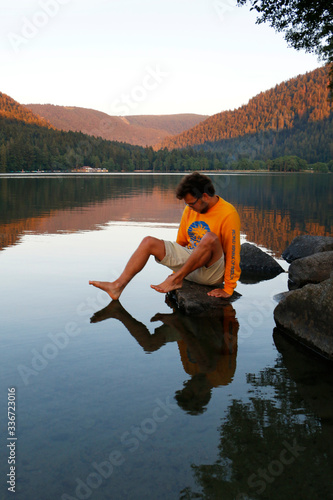 Male teenager touching his foot barely on the surface of alake while reflecting perfectly in the water