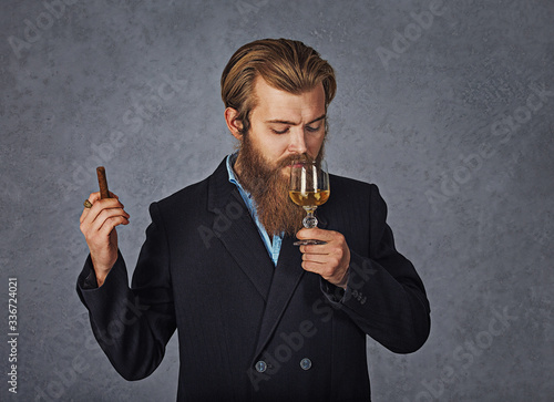 Young handsome bearded caucasian man sommelier tasting cognac with a cigar. Perfect skin and hairstyle. Wearing black suit on grey background. Studio portrait photo