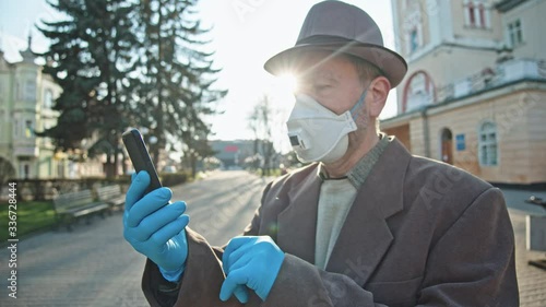 Quarantine, COVID-19. Old man with protection face mask and gloves using smartphone to communicate standing outside in the city. Pandemic. Urban lifestyle. photo