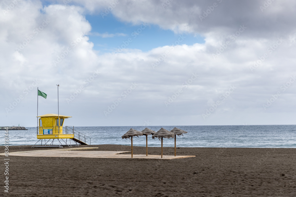 View of lifeguard tower on beach during windy sunny day with sea view and unique beach which has black sand on tower flutters green and yellow flag. View of lifeguard tower on beach during windy sunny day with sea view and unique beach which has black sand on tower flutters green and yellow flag.
