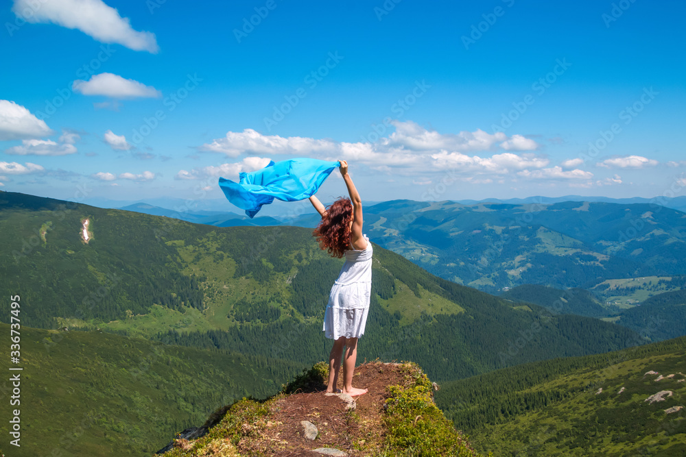 Obraz premium Young woman in white summer dress stands on a rock and looks into the valley, in her hands develops a lightweight blue scarf