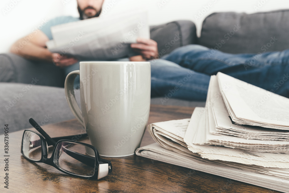 man relaxing on sofa reading a newspaper with stack of newspapers and coffee mug in foreground man relaxing on sofa reading a newspaper with stack of newspapers and coffee mug in foreground