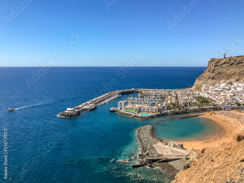 Fototapeta Naklejka Na Ścianę i Meble -  View from the air on the coast and bay in the city of Mogan on the Canary Islands where the bay harbor and people walking along the waterfront inside city