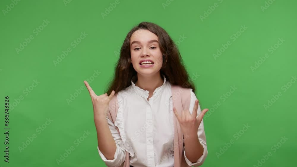Yeah, that's awesome! Overjoyed teenage brunette girl in white shirt showing rock and roll gesture, crazy hand sign, enjoying heavy metal music festival. studio shot isolated on green background Yeah, that's awesome! Overjoyed teenage brunette girl in white shirt showing rock and roll gesture, crazy hand sign, enjoying heavy metal music festival. studio shot isolated on green background