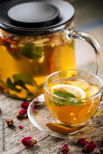 Closeup on the healthy herbal yellow apple tea with lemon and cinnamon in a glass teapot and mug on the wooden background decorated with roses, vertical