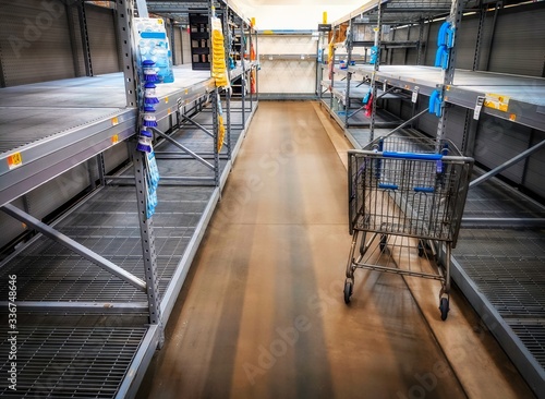 A view down a store shopping aisle showing empty shelves and a shopping cart.