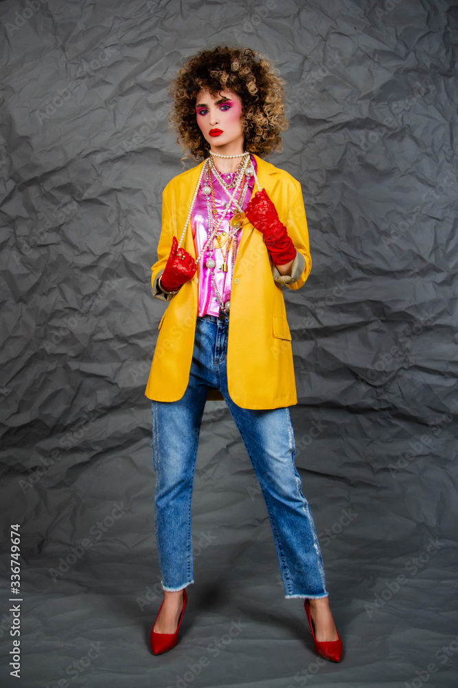 Girl in a yellow jacket and blue jeans with an afro hairstyle. Fashion of the eighties, disco era. Studio photo on gray background. Girl in a yellow jacket and blue jeans with an afro hairstyle. Fashion of the eighties, disco era. Studio photo on gray background.