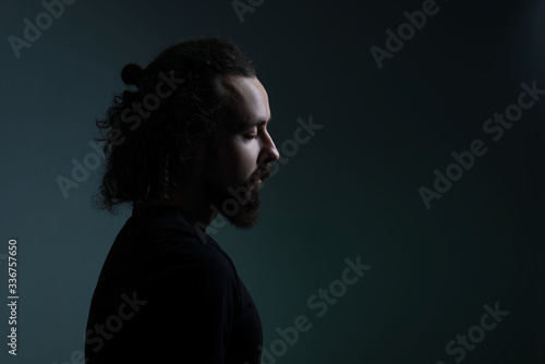 studio dramatic profile portrait of a young bearded guy of thirty years old, with long hair gathered, looks down. On a dark background. A ray of light on the face. Dramatic light