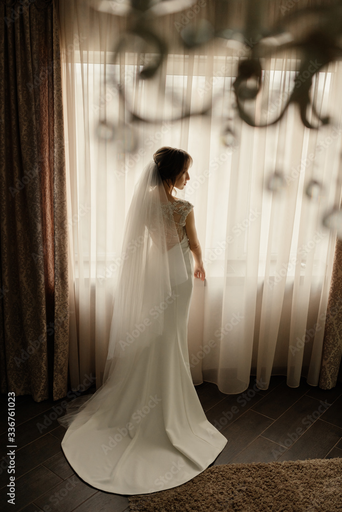 Full length elegant the bride stands in front of the window in a wedding dress at hotel. Wedding morning of the bride. Full length elegant the bride stands in front of the window in a wedding dress at hotel. Wedding morning of the bride.
