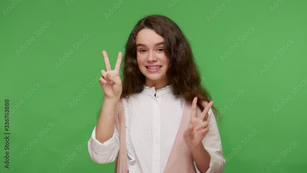 Optimistic teen brunette girl in white shirt showing victory, peace gesture with two fingers, positive about winning, celebrating success, triumph. indoor studio shot isolated on green background Optimistic teen brunette girl in white shirt showing victory, peace gesture with two fingers, positive about winning, celebrating success, triumph. indoor studio shot isolated on green background