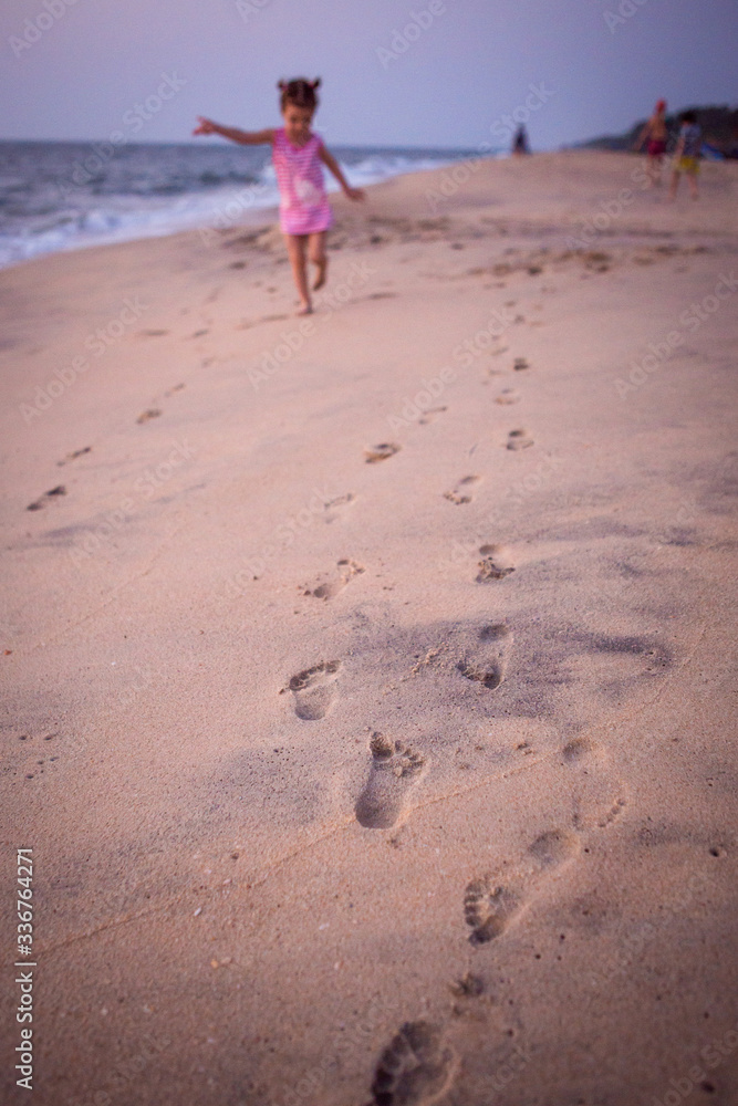 Footprints in the sand from children's feet. A girl in a pink t-shirt runs along the beach in the background. Footprints in the sand from children's feet. A girl in a pink t-shirt runs along the beach in the background.