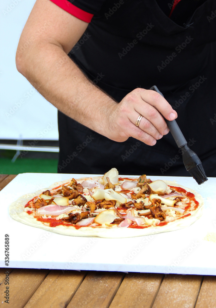 Italian cuisine. Preparation pizza with the hand of a chef. Dough, ketchup, bacon, mushrooms and cheese on a white board on a wooden table. Background image, copy space