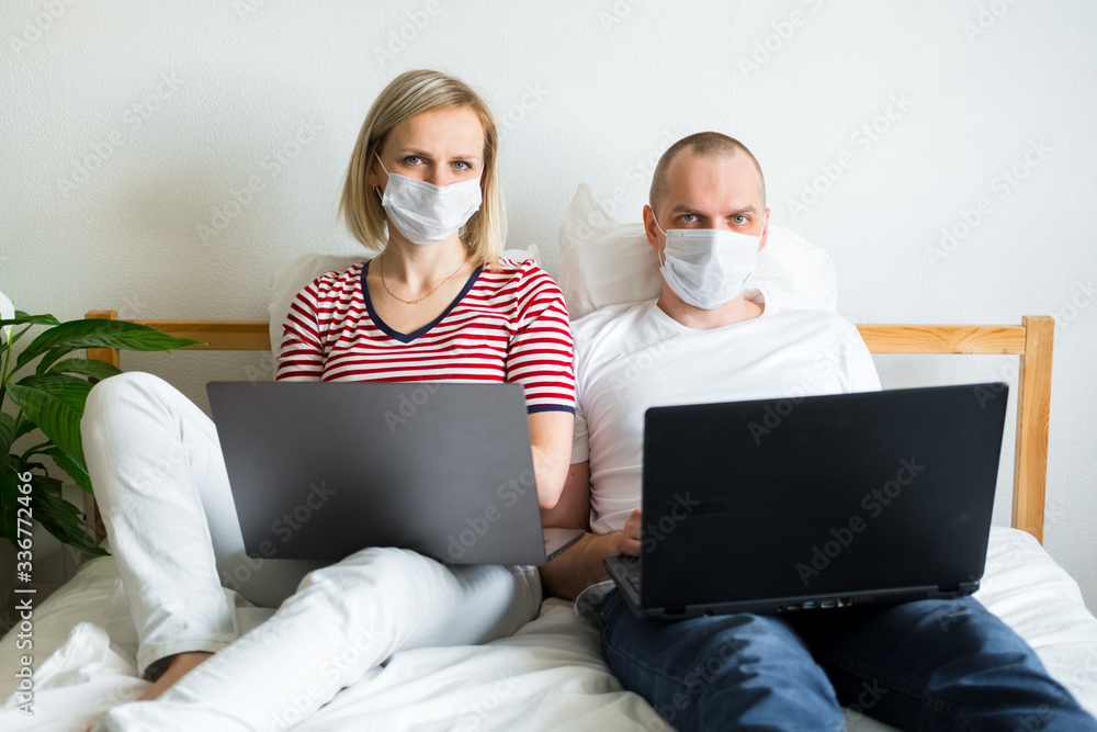 young family works remotely from home on bed at the computer. Quarantined couple coronavirus in medical masks. Stay home safe. Distance learning, education and work. Order food products online. young family works remotely from home on bed at the computer. Quarantined couple coronavirus in medical masks. Stay home safe. Distance learning, education and work. Order food products online.