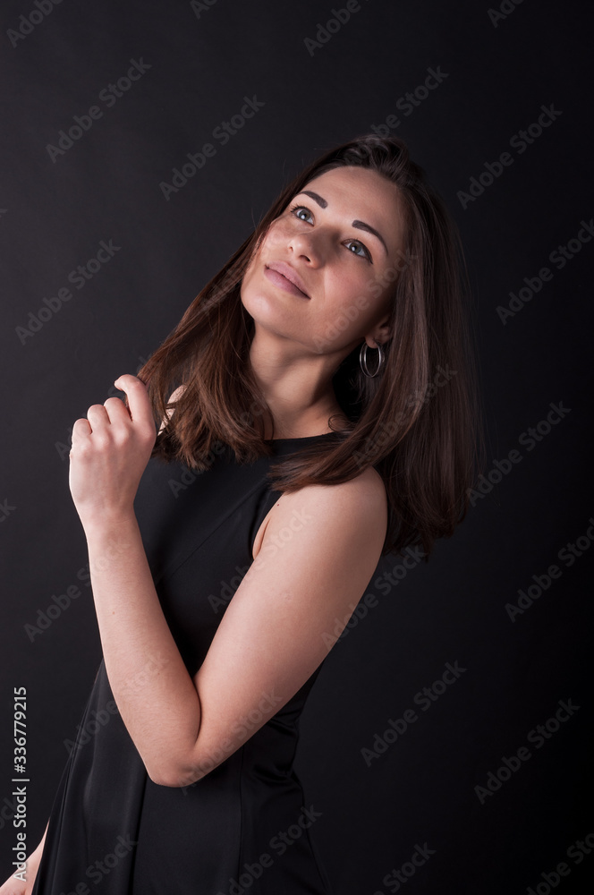 Satisfied young girl bowed her head and is touching her hair. Elegant black dress, so dark everywhere. Amazing girl with attractive appearance