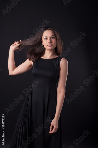 Beautiful young serious lady is touching her hair in the darkness and looking at the camera. Elegant black dress, so dark everywhere. Amazing girl with attractive appearance