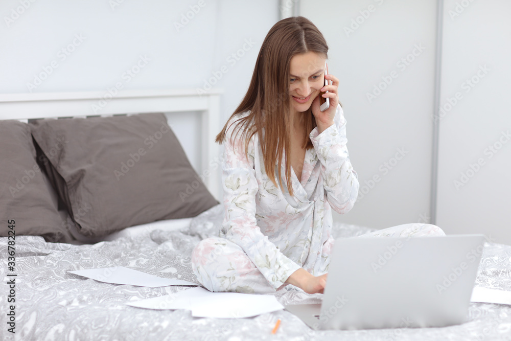 Attractive Young Woman Working from Home - Female Entrepreneur Sitting on Bed with Laptop Computer, Paperwork and tolking on the Cell Phone from Comfort of Home. quarantine. coronavirus Attractive Young Woman Working from Home - Female Entrepreneur Sitting on Bed with Laptop Computer, Paperwork and tolking on the Cell Phone from Comfort of Home. quarantine. coronavirus