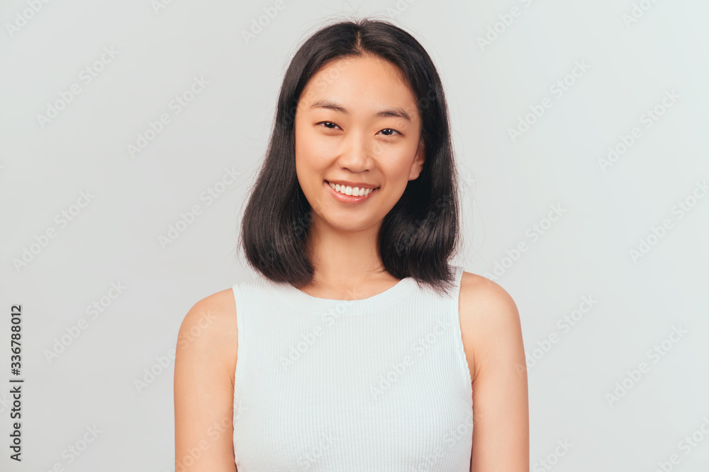 Portrait of girl tilts head and smiles sweetly showing white teeth. Beautiful young woman Asian appearance with black hair brown eyes stands isolated white background in Studio Portrait of girl tilts head and smiles sweetly showing white teeth. Beautiful young woman Asian appearance with black hair brown eyes stands isolated white background in Studio