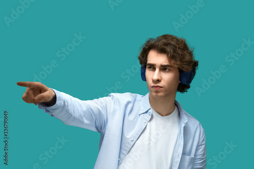 Portrait of a young beautiful man wearing white t-shirt and blue shirt in headphones point to something or somebody by his forefinger over blue background © 5M