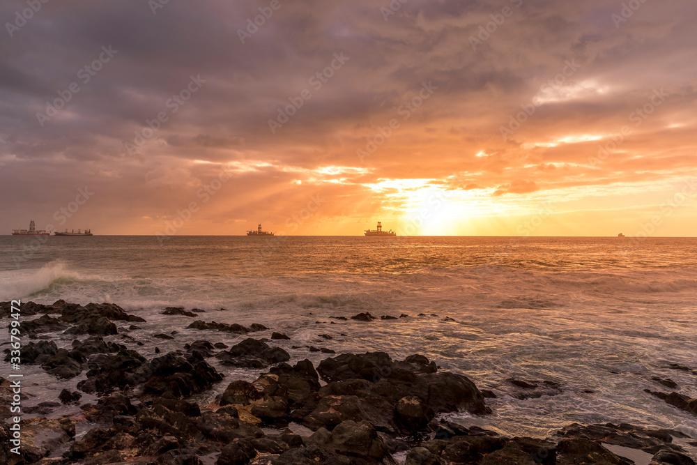 Fototapeta premium Sunrise from a stony shore to boats moored on the horizon with the rising sun and pesky shining through the clouds to the ocean with moving clouds from the Grand Canary Island.