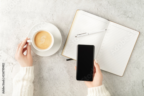 Beautiful women's hands holding up-to-date smartphone and writing in notepad with cup of coffee on concrete background. Women's business concept. Coffee break top view.